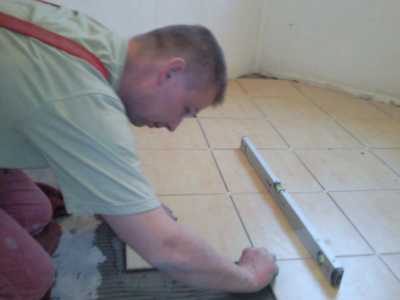 Pracownik w trakcie układania jasnych płytek podłogowych w pomieszczeniu w Rudzie Śląskiej, używający poziomicy do precyzyjnego montażu.